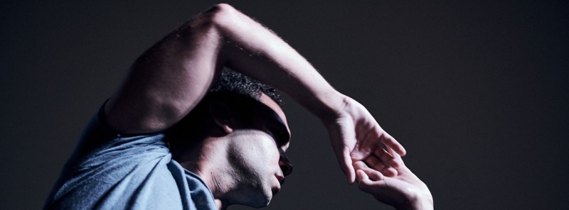 An image of a man with brown skin and black curly har. His right elbow is bent and facing up creating a shadow over his face.