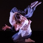 Two dancers dressed in white blouses and grey pants perform a lift. One dancer on their knees holds the other on their shoulder.