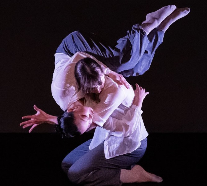 Two dancers dressed in white blouses and grey pants perform a lift. One dancer on their knees holds the other on their shoulder.