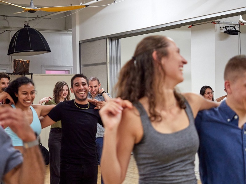 A picture of a dance class. There is a woman in front smiling with her arm around the shoulders of her dance partner.