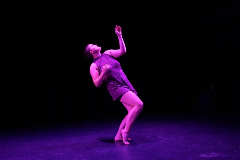 Rush, a white dancer in a purple dress, stands in a leaned back pose in purple lighting