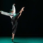 A dancer in a white top and black skirt performs a high kick on stage, with arms extended.