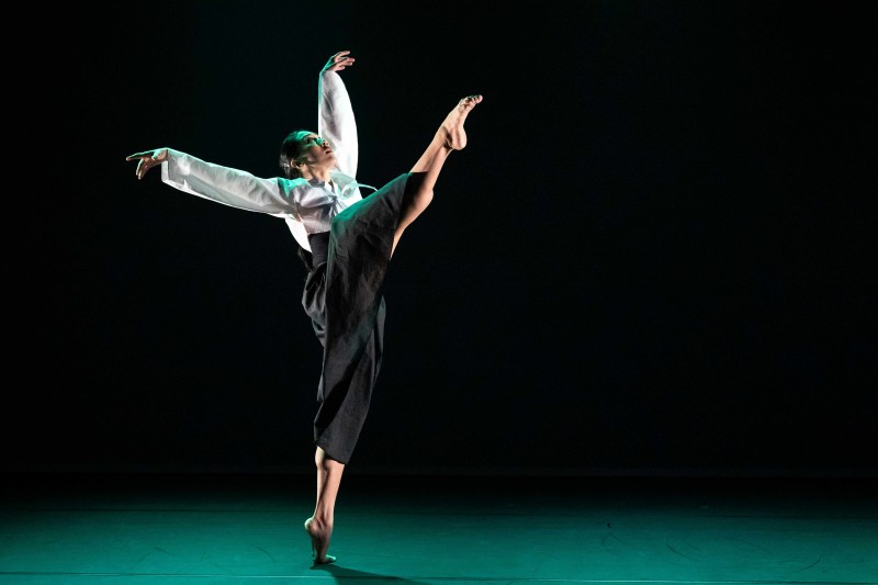 A dancer in a white top and black skirt performs a high kick on stage, with arms extended. 