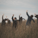Seven dancers stand in field of wheat up to their waists, arms outstretched