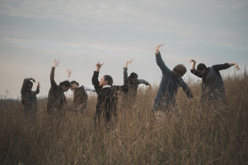 Seven dancers stand in field of wheat up to their waists, arms outstretched