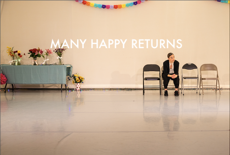 A woman sits in a folding chair with 3 empty chairs next to her. The room looks decorated for a party.