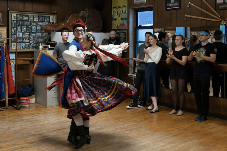 Two people dancing in Polish folk outfits in a dance workshop room with other dance workshop participants