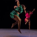 Two dancers jumping with their arms spread excitedly; one dressed in green and the other in pink