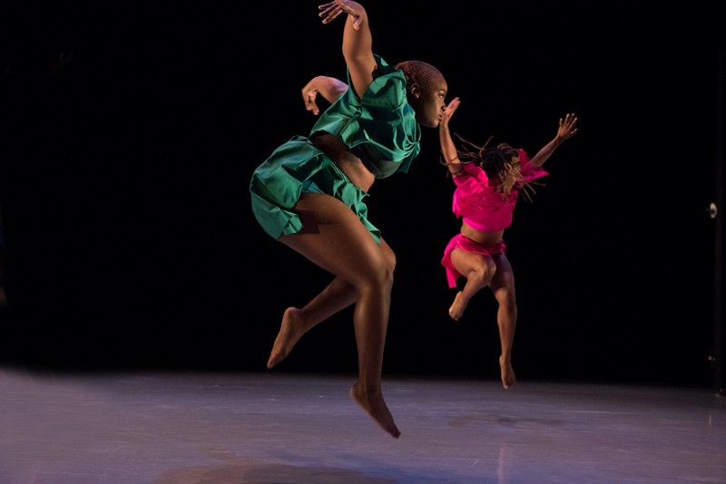 Two dancers jumping with their arms spread excitedly; one dressed in green and the other in pink