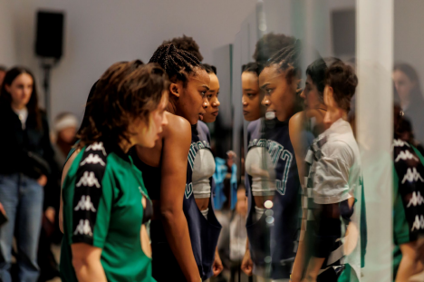 Two female identifying dancers looking at their reflection in a glass prop piece as part of a live art performance piece. 