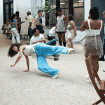 ID: People gathered in open courtyard. Dancer is in motion at a low level with a leg and an arm in the air.