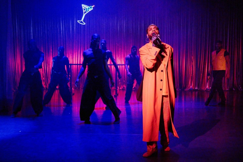 Brown hued man in red clothing singing into a microphone with dancers shadowed in the background 