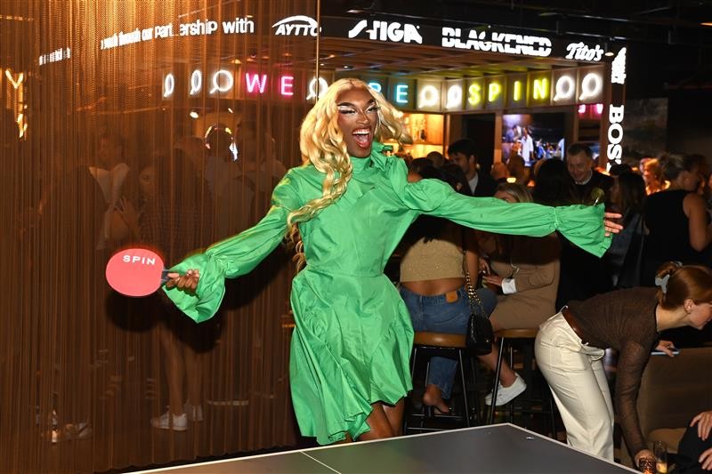 A person playing ping pong wearing a short longsleeved green dress. And there are people behind them talking