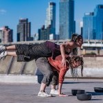 a woman in grey pants and purple top is balanced in a plank on the back of another dancer in an orange top city skyline behind
