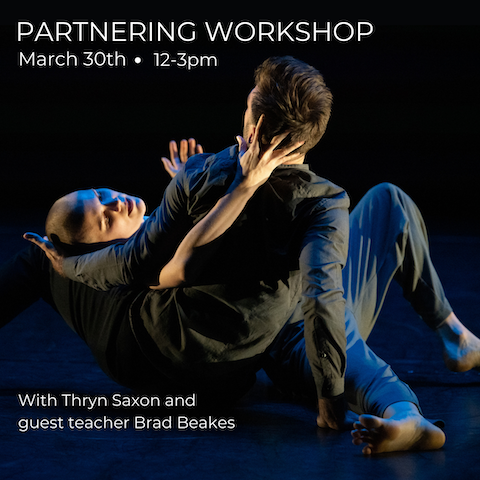 Two dancers lie in each others arms on a black marley floor with shadowed lighting. 