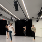 Four dancers stand together, twisting at the spine and looking over their left shoulders, right arms around at shoulder level.