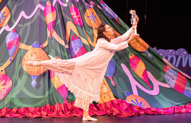A girl in a nightgown extends her leg and arms, admiring a nutcracker in front of a tree. 