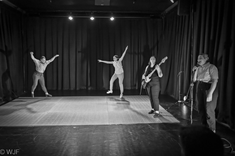 Two tap dancers in mid air, a bass player, and vocalist all performing on a small stage.