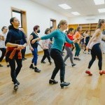 Tap dancers of many styles and ages flex their right foot on the floor in front of them while looking in the mirror.