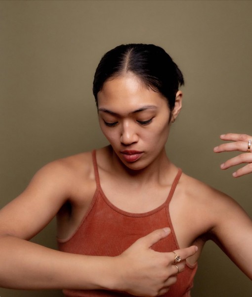 Keerati poses in front of a green background, her focus is inward and her arms are suspended around her torso.