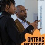 One Black man with glasses looks at a piece of art on a white wall with a young Black student.