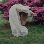 Woman in a white long dress lean back in a dance in front of pink blooming bush behind 