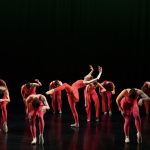Group of dancers wearing pink costumes and contracted over. Solo dancer in the center of the stage is in a high release.