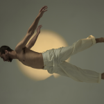 cis male jumping with his body horizontally to the floor with white pants on in front of a warm circular light