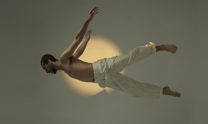 cis male jumping with his body horizontally to the floor with white pants on in front of a warm circular light