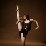 cis woman dressed in black skirt with brown shirt, with right leg up in front of a brown backdrop.