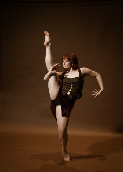 cis woman dressed in black skirt with brown shirt, with right leg up in front of a brown backdrop.