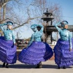 Three dancers in large purple hoopskirts and blue tops and hats dance outside on a sunny day in front of a fountain.