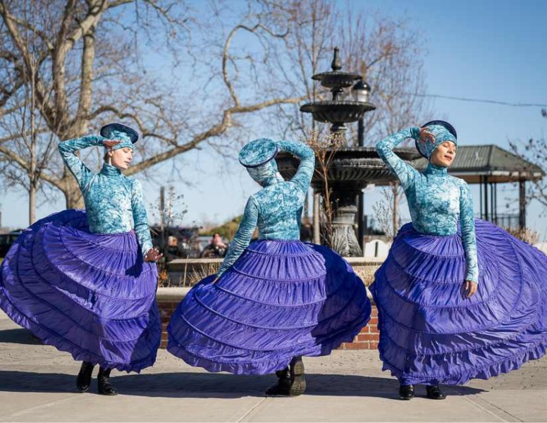 Three dancers in large purple hoopskirts and blue tops and hats dance outside on a sunny day in front of a fountain.