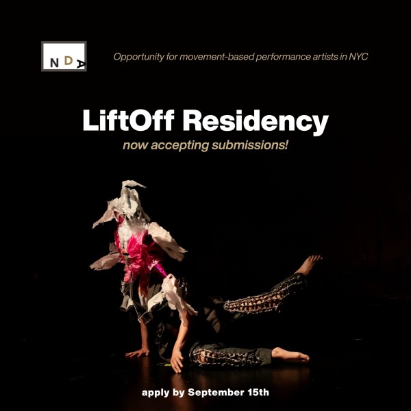 A performer sits on the floor against a dark background. She wears grey pants and a pink piñata on her head obscuring her face.