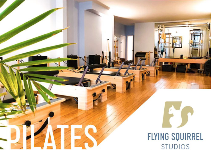 Pilates reformers lined up with Flying Squirrel Studios logo on the bottom right corner and greenery on the left side of the pic