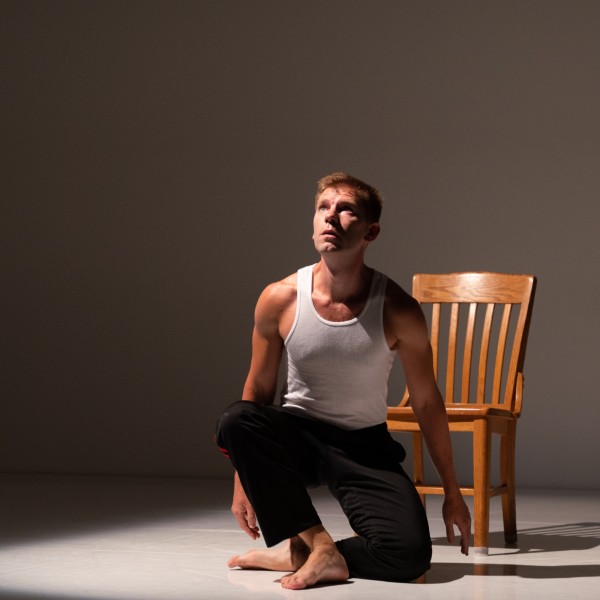 A man kneels on the floor in front of a wooden chair, looking upwards