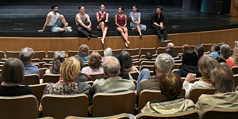 six dancers sit casually on stage in front of a small, seated audience. A dancer in the middle holds a microphone. 