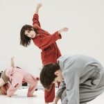 Three dancers in pink, red, & grey coveralls dance in a white space