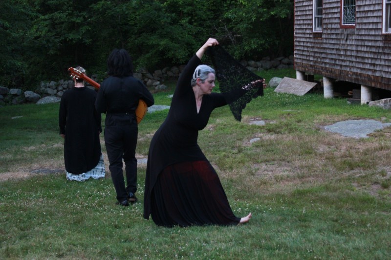 Dancer with a black lace shawl performing outside as two musicians stand nearby.