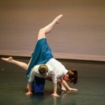 Two dancers in white tops and blue skirts perform a lift.