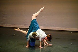 Two dancers in white tops and blue skirts perform a lift.