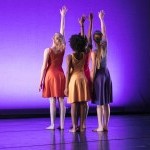 Four dancers with backs to camera. Right arm raised straight up. Dancers wearing knee length dresses of different colors. 