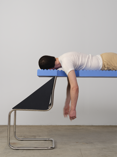 A photograph of a chair-like sculpture with a person reclining on it, facing away.