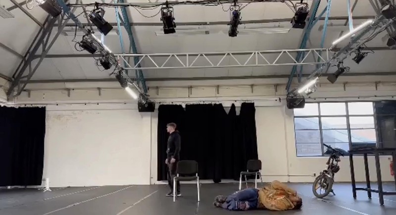 two performers in a rehearsal room. one is stood up while one is on the ground.