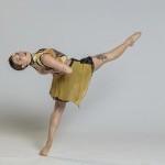 Dancer in mustard & black costume with bodysuit & small skirt with leg up and arms behind back.