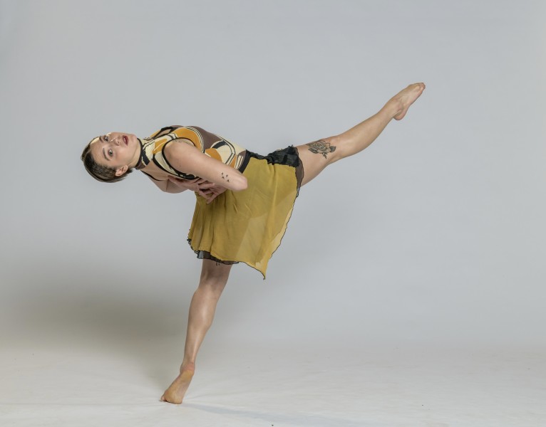 Dancer in mustard & black costume with bodysuit & small skirt with leg up and arms behind back.