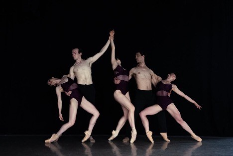 Five dancers posing in front of a black background. Three women pose on pointe, wearing skin-tone pointe shoes and purple leotar