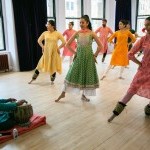 A picture o several dances dressed in colorful tunics in a kathak pose with a tabla artist sitting in front of them