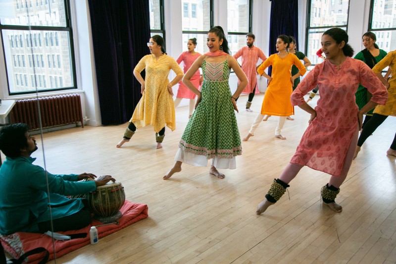 A picture o several dances dressed in colorful tunics in a kathak pose with a tabla artist sitting in front of them 