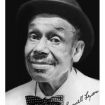Black and white headshot of a man wearing a white shirt, polka dotted bow tie, and black hat with his signature in black.